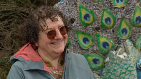 Trudie has curly brown hair and wears sunglasses. She is standing beside the peacock that Curley made for her mother.