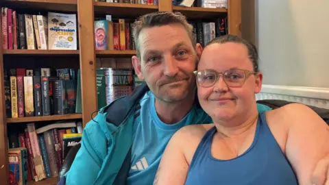 Mark and Elen take a selfie on a sofa with a bookcase full of books behind them. Elen has her hair tied back and is wearing glasses and a blue vest top, and Mark has short hair and stubble and is wearing a blue jacket and blue Addidas t-shirt. 
