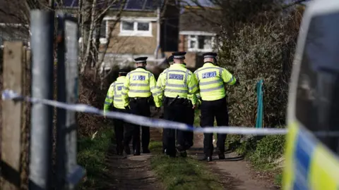 BBC A group of uniformed Sussex Police officers walk down a lane with their backs to us. In the foreground the edge a police van can be seen on the right hand side. The land is cordoned off with police tape.