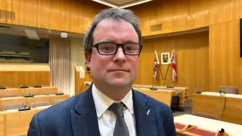 Martin Heath/BBC Henry Vann with short dark hair wearing glasses, a blue jacket, white shirt and grey tie, standing in a council chamber made up of wooden desks with seats behind, each bearing a microphone on a small plastic unit. A portrait of the King and two union flags can be seen on the wall in the background.