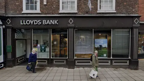 The exterior of Lloyds Bank in Horncastle. The high street building is painted black and there are two people, with their faces blurred, walking in front of the building. 