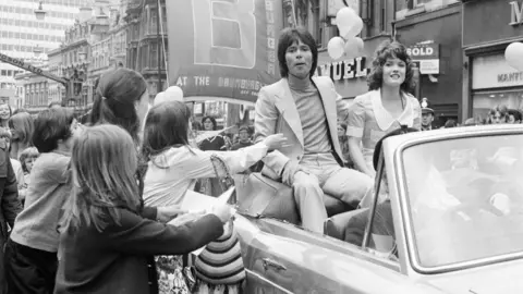 Getty Images Cliff Richard and Deborah Watling in an open top car with crowds of people asking for autographs