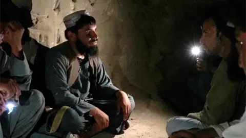 A bearded man in a traditional dark tunic and trousers, wearing a brimless hat, sits cross-legged on the bare floor of a dark cave. He is on the left of the picture and is looking at a man sitting on the right, who is holding a torch.