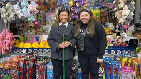 Two women pose in front of a stall of toys, including swords, rubber ducks, and cuddly animals.