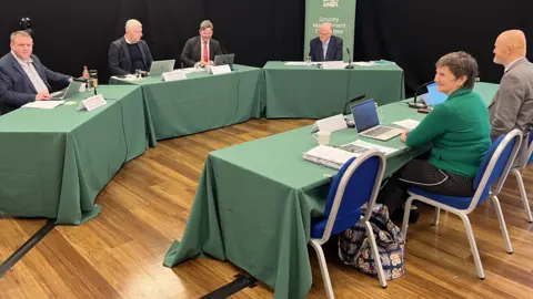 A group of four people sitting behind a long table with a green tablecloth. Facing them are a man and a woman facing the table, again with a green table cloth on it. 