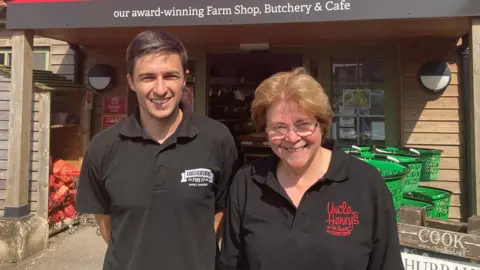 Sam with short brown hair and Meryl Ward with short hair and glasses both wearing black polo shirts and smiling in front of their farm shop