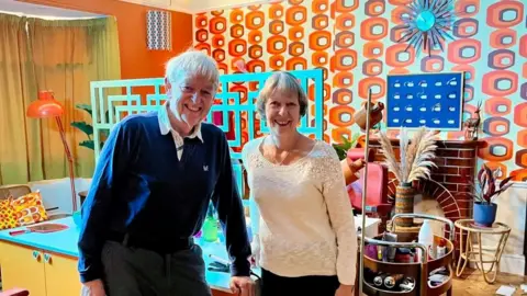 Mark Cargill A man and a woman, both with grey hair, stand in front of the set of a hairdressing salon with bright orange wallpaper and a bright blue table.