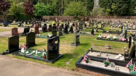Rows of burial plots, most with black gravestones, set on a lawn, with a wooden fence and well-established trees in the background.