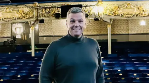 The Tyne Theatre & Opera House Jonathan Higgins, with short grey hair, wearing a grey long-sleeve top and black pants and trainers, has his hands in his pockets as he stands on a stage. There are seats behind him.