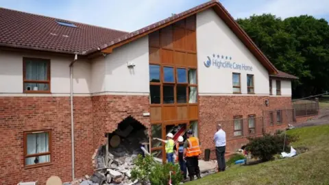 PA Media Damaged front of the Highcliffe Care Home in Sunderland which has a large hole in the front of the building. A large amount of debris can be seen inside the hole. The building is large with the bottom made out of red brick. The first floor of the building is painted white and has a pointed roof. Three people in hard hats and hi-vis can be seen next to the hole along with a man in a shit.