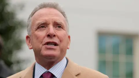Steve Witkoff in striped shirt and red tie, wearing a brown coat