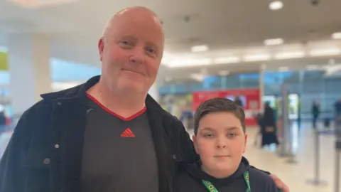 A man is stood in the airport wearing a black jacket with his arm around his son, who is also wearing black. They are looking directly at the camera.