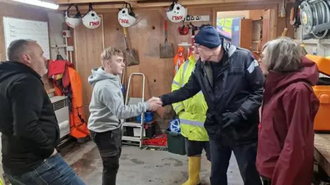 RNLI Rob Beckley is saking the hand of a young man. They are standing with three other people in a shed filled with rescue equipment.