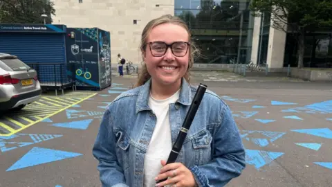 A smiling woman stands in front of the Ulster University Belfast building holding a flute