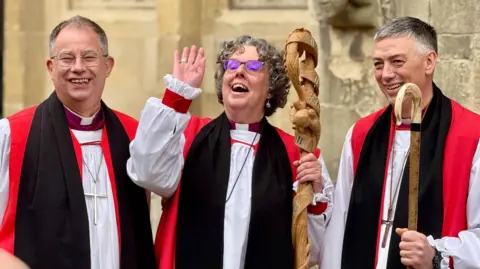 James Wood Bishop Gregory in the middle of two other bishops  - everyone is in white, red and black robes and smiling at the camera. Bishop Gregory is waving.