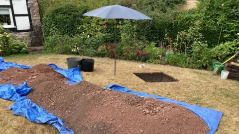 A mound of earth has been dug up in someone's garden. The earth has been emptied onto a blue sheet of tarpaulin with a deep square-shaped hole dug in the corner of the land.