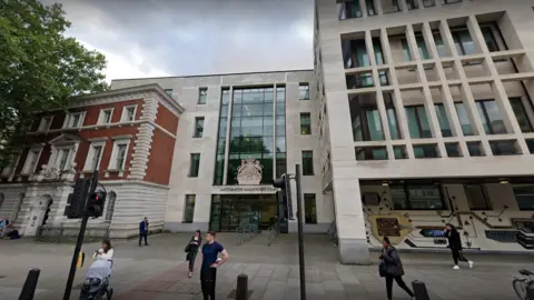 An exterior view of Westminster Magistrates' Court. People can be seen walking past the glass and beige stone building which also has a coat of arms above the front doors.