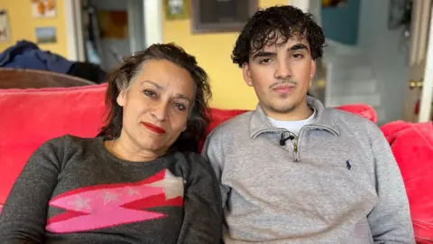 Seb and his mother sit side by side on a couch and look at the camera. She is wearing a grey jumper with a pink shooting star on it and has long dark hair pulled back off her face, while Seb is wearing a light grey jumper.