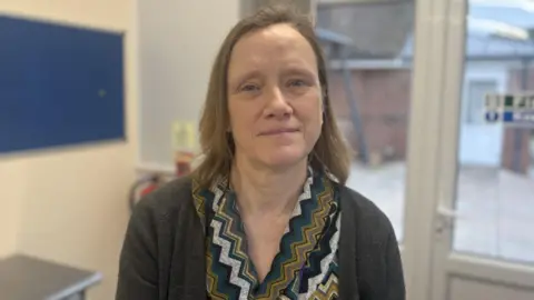 A woman with light brown, shoulder-length hair is sitting in a medical setting. She is wearing a grey cardigan and zig-zag patterned top.