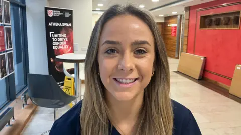 Georgia Ross is standing in a brightly lit hallway. She has blonde hair, is smiling and is wearing a navy top. Chairs and a table can be seen behind her.