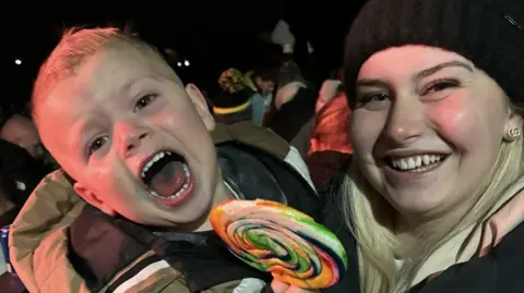 Sophie smiling widely with her boy, Remi, three, in her arms. He is also smiling and possibly shouting, with a big lollipop in his hand.