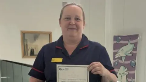 A woman in uniform is holding a piece of paper and looking at the camera. She is standing in a corridor.
