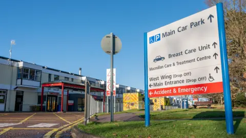 Martin Barber/BBC Outside the Queen Elizabeth Hospital in King's Lynn. A sign can be seen that is signposting people to various places, including the main car park, breast care unit, Macmillan Care and Treatment Centre, the West Wing, main entrance, and accident and emergency departments. Two yellow ambulances can also be seen.