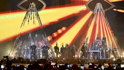 Getty Images The Pet Shop Boys performing a show, with bright lights shining down on them and musicians in silver costumes behind.