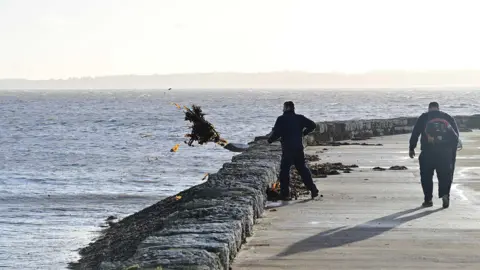 Pacemaker Two men in dark clothing clear a sea wall of debris following a storm. 