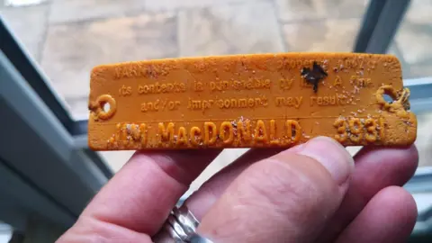 Nikki Banfield/ BareFoot Photographer An orange tag with the name 'Jim MacDonald' on it. Nikki is holding it in her hand in front of a window. 