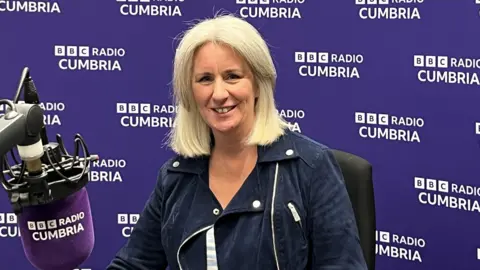 Heather Wainwright sitting in a BBC Radio Cumbria studio, with a branded purple microphone in front of her and a branded purple wall behind her. She has blonde/white hair in a bob and is wearing a blue jacket. She is smiling at the camera.