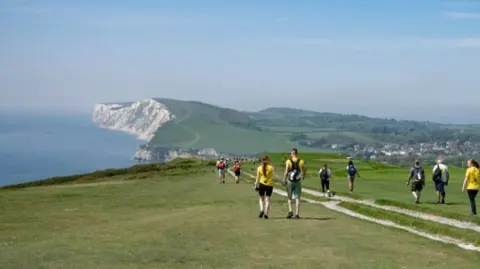 Walk the Wight A scenic shot of the Isle of Wight coast, with competitors following a trail, many carrying backpacks, as they head towards the cliffs in the distance.