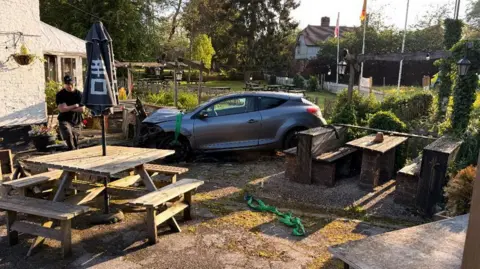 A car in a pub garden with damage to the front of the vehicle and the pub garden furniture.