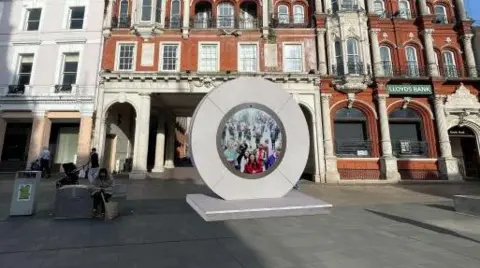 A computer generated image of what the portal could look like. It is a grey circle with a screen in the middle, it is on the Cornhill in Ipswich with buildings in the background. 