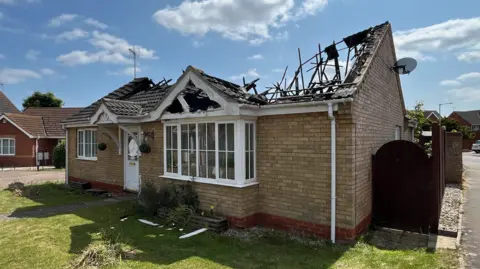 A view of the front of a bungalow in a residential street. Its damaged roof can be seen badly damaged and caved in.  