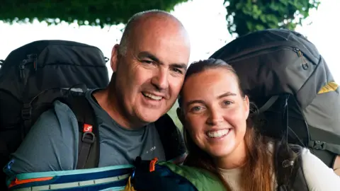 BBC/Studio Lambert Andrew and Molly. Andrew is bald, wearing a grey t-shirt and carrying rucksacks on his front and back. Molly has red hair tied back wearing a white t-shirt and carrying a large rucksack on her back.