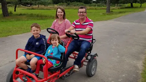 Darren Wheatley Nicola, Darren and their two children on a pedal-powered cart red cart. Nicola is wearing a red gingham dress and Darren is wearing a pink and white striped polo shirt, jeans and trainers. Ffion is wearing a blue cardigan and has her finger in her mouth. Oscar is wearing a navy hoody and shorts. All are smiling at the camera. 