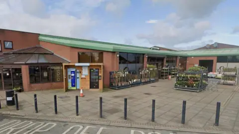 The exterior of a supermarket building with mainly a flat roof, with green panelling. There are bollards separating a drop off zone from the front of the store and there are rows of plants for sale outside.
