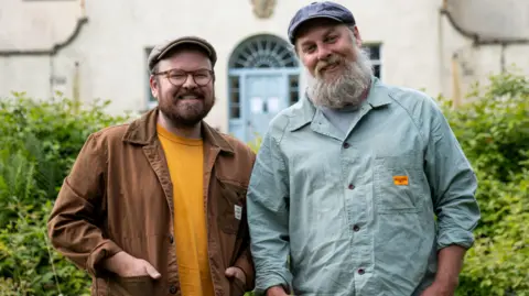 Shelley Richmond/Hello Halo/BBC Scotland Banjo Beale - a man with glasses, a flat cap and a beard, wearing a brown jacket and orange shirt and Ro Christopher, a man in a aquamarine shirt, with a white beard and a flat cap.