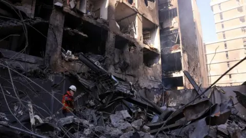 A rescue worker wearing a helmet and protective gear searches through rubble inside a heavily damaged multi-storey building with collapsed walls, twisted metal and debris.