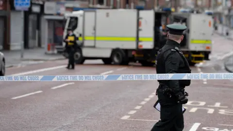 Getty Images A police cordon has been placed along a street in Belfast city centre. A male police officer in full uniform is standing beside the cordon but looking away from the camera. A female police officer is standing beside a white van which is concealed behind the cordon. 