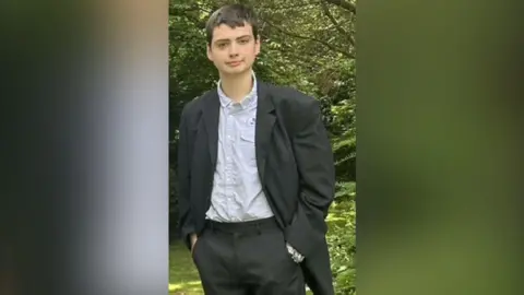 A young man with short black hair, wearing a grey shirt, black jacket, and black trousers, stands in front of a tree with his hands in his pockets.
