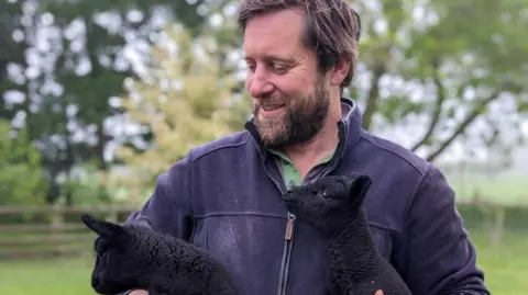 Tom Martin is stood outside in a field holding two black lambs in each arm. He wears a blue zip up fleece with a green shirt underneath and had dark brown hair and a matching colour beard. He is looking at the lambs and smiling. 