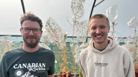 Two men are stood inside a plastic greenhouse in front of dying lettuce plants that have bags over the top of them. The man on the left is wearing glasses and a green t-shirt with white writing, he has brown hair and a beard. The man on the right has short blond hair and is wearing a grey hoodie with black writing. 