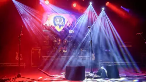 A stage set with drums, microphones, speakers and lit in red and blue. A sign at the back reads UNDER 18S BATTLE OF THE BANDS.