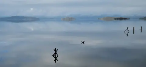 Judith Shaw A calm, glassy lake reflecting a grey sky and distant low hills. A few weathered wooden posts and cross‑shaped markers rise from the water in the foreground.