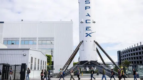 A row of people walk past a large white rocket with blue "space x" logo written on it.