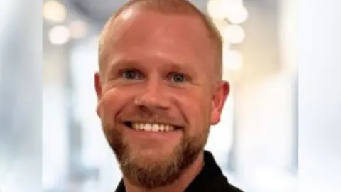 A man with a shaved head and beard smiles at the camera 