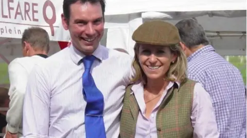 James Ewart Racing A man in a shirt and blue tie has his arm around a woman in a cap with a checked gilet over a lilac blouse