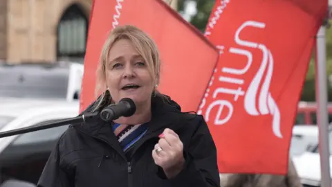 PA Media Sharon Graham, General Secretary of the Unite union, pictured at a previous trade union event.  She has blonde hair tied back from her face and is wearing a multicoloured top or dress under a black rain coat.  Red and white Unite flags are flying behind her. She is speaking into a microphone and holding one closed fist in front of her. 